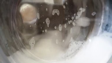 Beyaz çamaşır makinesi çamaşır yıkar. Dönen davul çamaşır makinesinin görüntülerini kapat. Kalın köpük, su ve kabarcıklar çamaşır makinesinin cam kapısından görülebilir. Santrifüj döndürmesi