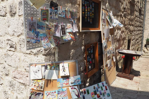 Kotor, Montenegro, August 11, 2022: souvenir cards, drawings, pictures and magnets are hung on the wall and on a metal sign. Tourist walks around the old town. Sea summer breeze. Ancient stone walls