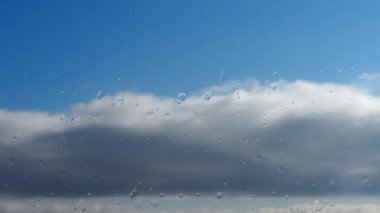 Sisli camlara yağmur damlaları. Mavi gökyüzünde yatay kabarık bulutlar uçar. Bulutların gerçek zamanlı hareketi. Yan taraftan güneş ışığı az. Işık buharı ve nem