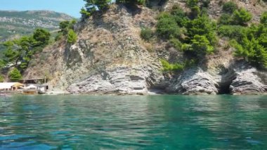 Petrovac, Karadağ 07.22.24 Tatilciler, güneşlenenler ve yüzücülerle kıyıdan geçen bir tekne. Oteller, güneş şemsiyeleri, Adriyatik dalgaları, şamandıralar. Plaj turizmi. Tatil seyahati. Yaz günü. Kumlu çakıl.