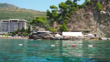 Petrovac, Karadağ 07.22.24 Tatilciler, güneşlenenler ve yüzücülerle kıyıdan geçen bir tekne. Oteller, güneş şemsiyeleri, Adriyatik dalgaları, şamandıralar. Plaj turizmi. Tatil seyahati. Yaz günü. Kumlu çakıl.