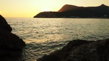 Dalgalar sersemletici günbatımında yavaşça kıyıya vurur. Altın güneş ışığı, dalgaların kıyıya vuruşunu yansıtır. Gün batımında deniz kenarında sakin bir atmosfer yaratır. Karadağ yazı, Canj