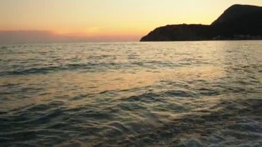 Dalgalar sersemletici günbatımında yavaşça kıyıya vurur. Altın güneş ışığı, dalgaların kıyıya vuruşunu yansıtır. Gün batımında deniz kenarında sakin bir atmosfer yaratır. Karadağ yazı, Canj