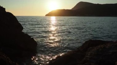 Dalgalar sersemletici günbatımında yavaşça kıyıya vurur. Altın güneş ışığı, dalgaların kıyıya vuruşunu yansıtır. Gün batımında deniz kenarında sakin bir atmosfer yaratır. Karadağ yazı, Canj