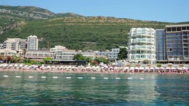 Petrovac, Karadağ 07.22.24 Tatilciler, güneşlenenler ve yüzücülerle kıyıdan geçen bir tekne. Oteller, güneş şemsiyeleri, Adriyatik dalgaları, şamandıralar. Plaj turizmi. Tatil seyahati. Yaz günü. Kumlu çakıl.