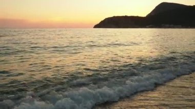 Dalgalar sersemletici günbatımında yavaşça kıyıya vurur. Altın güneş ışığı, dalgaların kıyıya vuruşunu yansıtır. Gün batımında deniz kenarında sakin bir atmosfer yaratır. Karadağ yazı, Canj.