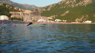 Canj Montenegro 07.20.24 Bir tekne ve tatilciler, güneşlenenler ve yüzücülerle dolu bir sahil. Oteller, güneş şemsiyeleri, Adriyatik dalgaları, şamandıralar. Plaj turizmi. Tatil seyahati. Yaz günü. Çakıl taşı plajı.