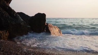 Dalgalar günbatımında okyanusta yavaşça kayalık kıyılarda tur atar. Altın güneş ışığı, dalgaların kıyıya vuruşunu yansıtır. Gün batımında deniz kenarında sakin bir atmosfer yaratır. Karadağ yazı, Canj.
