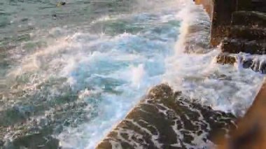 Dalgalar sersemletici günbatımında kayalık sahilde nazikçe tur atar. Güneş ışığı dalgaları yansıtıyor. Kıyı iskelesine köpükle yuvarlanarak deniz yoluyla sakin bir atmosfer yaratıyor. Karadağ yazı, Sutomore