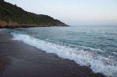 Dalgalar sersemletici günbatımında yavaşça kıyıya vurur. Güneş ışığı, dalgaları kıyıya vururken yansıtır. Gün batımında deniz kenarında sakin bir atmosfer yaratır. Karadağ yazı. Mavi saat.