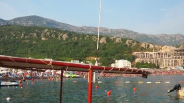 Canj, Karadağ 07.20.2024 Boat ve tatilciler, güneşlenenler ve yüzücülerle dolu bir sahil. Oteller, güneş şemsiyeleri, Adriyatik dalgaları, şamandıralar. Plaj turizmi. Tatil seyahati. Yaz günü. Çakıl taşı plajı.