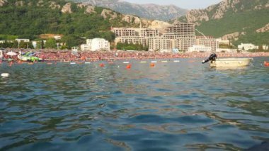 Canj, Karadağ 07.20.2024 Boat ve tatilciler, güneşlenenler ve yüzücülerle dolu bir sahil. Oteller, güneş şemsiyeleri, Adriyatik dalgaları, şamandıralar. Plaj turizmi. Tatil seyahati. Yaz günü. Çakıl taşı plajı.