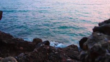 Köpüklü dalgalar mavi saatte kayalık kıyıya çarpar. Güneş ışığı dalgalardan yansıyarak kayaların üzerine yuvarlanır ve deniz kenarında pembe gün batımında sakin bir atmosfer yaratır. Karadağ yazı