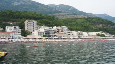 Sutomore, Karadağ 07.21.24 Boat ve tatilciler, güneşlenenler ve yüzücülerle dolu bir sahil. Oteller, güneş şemsiyeleri, Adriyatik dalgaları, şamandıralar. Plaj turizmi. Tatil seyahati. Yaz günü. Çakıl taşı plajı.