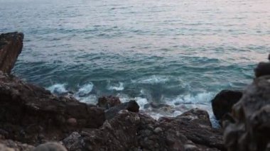 Köpüklü dalgalar mavi saatte kayalık kıyıya çarpar. Güneş ışığı dalgalardan yansıyarak kayaların üzerine yuvarlanır ve deniz kenarında pembe gün batımında sakin bir atmosfer yaratır. Karadağ yazı
