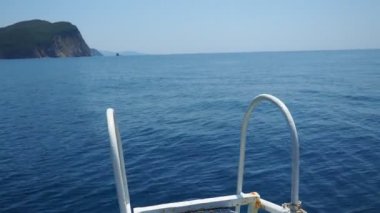 Durgun sular, berrak mavi gökyüzünün altında sonsuza dek uzanıyor. Tekne ön planındaki gösteriler ayrıntıları yıpratırken, uzak tepeler sakin bir zemin oluşturuyor. RT Dubovica Buljarica, Svetac Rock, Karadağ yakınlarındaki Kayalıkları