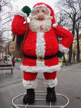 Sremska Mitrovica, Sırbistan 24,12,2024: Parlak kırmızı ve beyaz süslemelerle süslenmiş büyük bir Noel Baba figürü açık havada elinde bir çanta ve şeker kamışı tutarak duruyor. Yağmurlu bir atmosferde neşeli bir ruh.