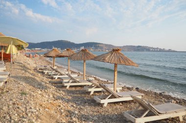 Bar, Karadağ: 07.20.2024 Güneş şezlongları ve sazdan yapılmış şemsiyeler, ziyaretçilerin deniz kenarında sakin bir günün keyfini çıkardıkları Bar Beach 'te sergileniyor. Yumuşak dalgalar kayalık sahilde tur atar. Akdeniz.