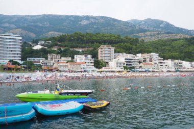 Sutomore Montenegro 07.20.24 Renkli botlar güneşlenenlerle dolu, hareketli bir plaj yakınlarında berrak sularda yüzüyor. Etrafı yemyeşil dağlarla çevrili sahil kasabası canlı yaz titreşimleri sunuyor. Adriyatik Denizi.