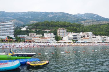 Sutomore Montenegro 07.20.24 Renkli botlar güneşlenenlerle dolu, hareketli bir plaj yakınlarında berrak sularda yüzüyor. Etrafı yemyeşil dağlarla çevrili sahil kasabası canlı yaz titreşimleri sunuyor. Adriyatik Denizi.