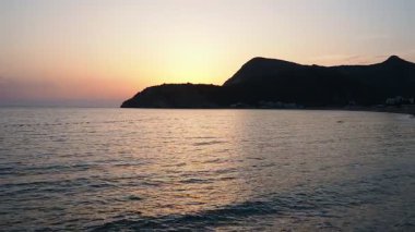 Dalgalar sersemletici günbatımında yavaşça kıyıya vurur. Güneş ışığı, dalgaları kıyıya vururken yansıtır. Gün batımında deniz kenarında sakin bir atmosfer yaratır. Karadağ yazı. Canj, Bar Bölgesi.
