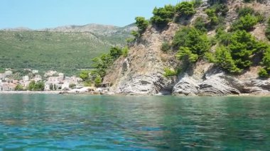 Petrovac, Karadağ 07.22.24 Tatilciler, güneşlenenler ve yüzücülerle kıyıdan geçen bir tekne. Oteller, güneş şemsiyeleri, Adriyatik dalgaları, şamandıralar. Plaj turizmi. Tatil seyahati. Yaz günü. Kumlu çakıl.