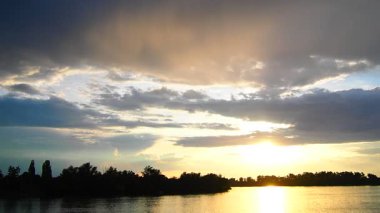 Alacakaranlıkta yemyeşil ağaçlar ve dinamik bulut oluşumlarıyla çevrili huzurlu bir göl üzerinde ışıl ışıl parlayan bir halo güneşinin büyüleyici güzelliğini deneyimleyin. Doğa sihrini ortaya çıkarır. River Sava, Sırbistan.