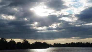 Parlak ışık demetleri bulutları delip geçerken huzurlu bir nehir manzarasının dönüşümüne tanık olun. Bu büyüleyici zaman atlaması doğanın güzelliğini büyülü bir şekilde yansıtıyor..