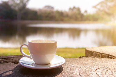 Beyaz bir fincan sıcak espresso kahve fincanı ahşap bir zemine ve güneş ışığı arkaplanlı doğal bir havuza yerleştirilmiş, kahve sabahı