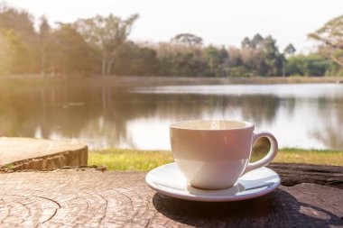 Beyaz bir fincan sıcak espresso kahve fincanı ahşap bir zemine ve güneş ışığı arkaplanlı doğal bir havuza yerleştirilmiş, kahve sabahı