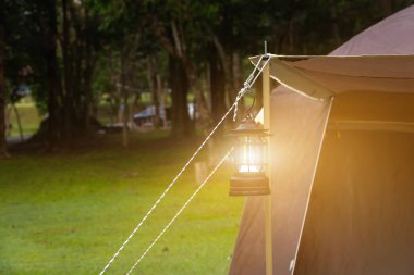Açık havada yemyeşil bir tentenin altında asılı siyah bir kamp fenerinin yakın çekimi. LED fener bir iple asılmış. Arka planda ağaçlar ve çimenli homurdanmalar görünüyor