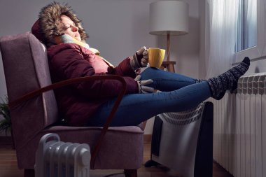 Woman in jacket, scarf and hand gloves indoors on a chilly winter day, energy and gas crisis, cold room, heating problems.