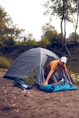 Man camping in nature, setting up the tent for overnight staying near forest river.