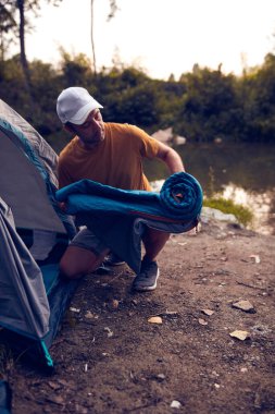 Man camping in nature, setting up the tent for overnight staying near forest river.