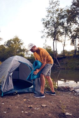 Man camping in nature, setting up the tent for overnight staying near forest river.