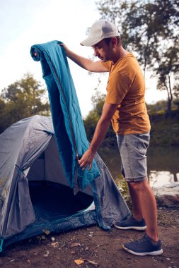Man camping in nature, setting up the tent for overnight staying near forest river.