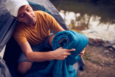 Man camping in nature, setting up the tent for overnight staying near forest river.
