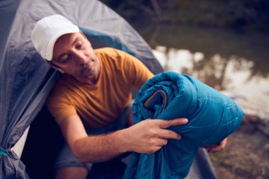 Man camping in nature, setting up the tent for overnight staying near forest river.