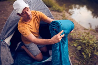 Man camping in nature, setting up the tent for overnight staying near forest river.