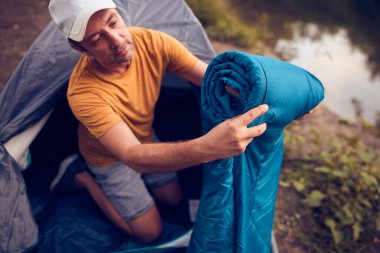 Man camping in nature, setting up the tent for overnight staying near forest river.