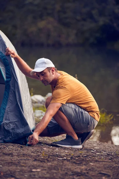 Man camping in nature, setting up the tent for overnight staying near forest river.