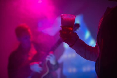 Elinde plastik bira bardağı olan ve arka planda gitar çalan bir kadın..