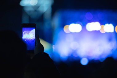 Elinde akıllı telefon ve sahne ışıkları olan konser kalabalığının siluetlerini tutan kişi