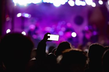 Elinde akıllı telefon ve sahne ışıkları olan konser kalabalığının siluetlerini tutan kişi