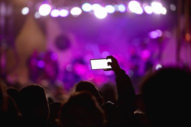 Elinde akıllı telefon ve sahne ışıkları olan konser kalabalığının siluetlerini tutan kişi