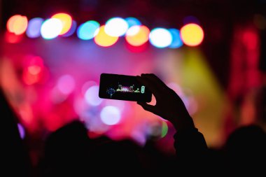 Elinde akıllı telefon ve sahne ışıkları olan konser kalabalığının siluetlerini tutan kişi