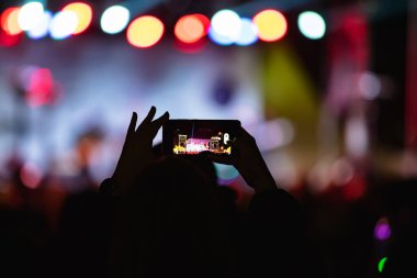 Elinde akıllı telefon ve sahne ışıkları olan konser kalabalığının siluetlerini tutan kişi