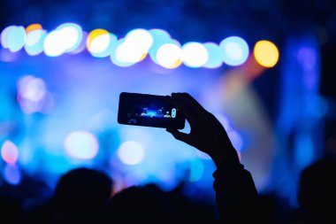 Elinde akıllı telefon ve sahne ışıkları olan konser kalabalığının siluetlerini tutan kişi