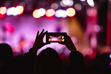 Elinde akıllı telefon ve sahne ışıkları olan konser kalabalığının siluetlerini tutan kişi
