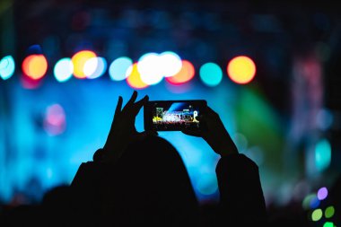 Elinde akıllı telefon ve sahne ışıkları olan konser kalabalığının siluetlerini tutan kişi